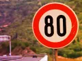 Segnale di limite massimo nei centri abitati: 80 km/h. Strada panoramica con vegetazione sullo sfondo.