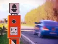 Autovelox arancione sul ciglio della strada, pronto a multare chi sfora la velocità. Un'auto blu sfreccia via, ignara.