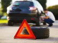 Triangolo di emergenza e ruota sgonfia accanto a un'auto nera, con un uomo che lavora alla ruota posteriore.