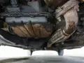 Sotto un'auto, si nota una **perdita olio motore dalla testata**, con accumuli scuri e gocciolamenti sul carter e sul tubo di scarico.