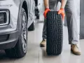 Meccanico con guanti arancioni tiene un nuovo pneumatico, pronto per essere montato su un'auto grigia. La durata gomme auto è fondamentale per la sicurezza.