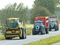 Tre trattori, uno giallo e due blu con rimorchi, percorrono una strada. Si rispettano i limiti di velocità macchine agricole.