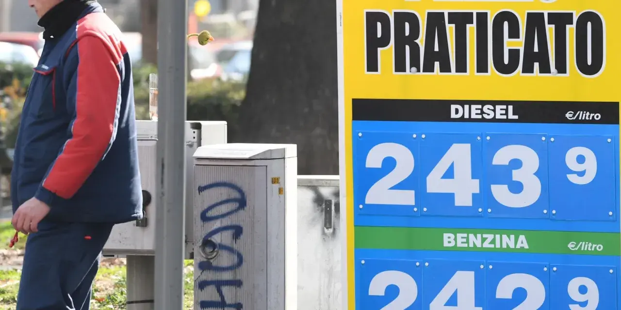 Prezzo praticato per la benzina: 2,429 €/litro. Un uomo in divisa osserva il cartello.