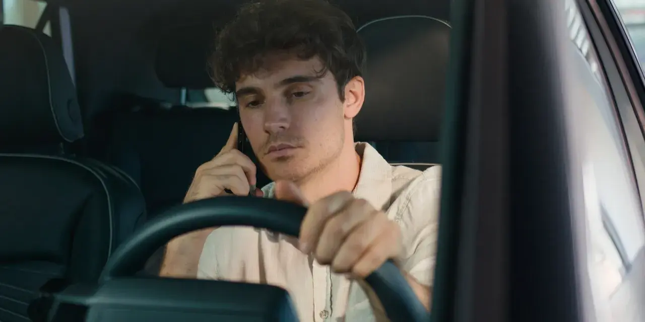 Giovane uomo al volante con il cellulare all'orecchio, distratto dalla conversazione.