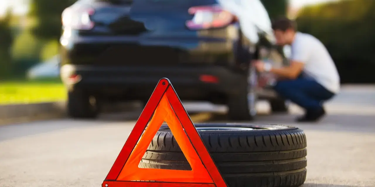 Triangolo di emergenza e ruota sgonfia accanto a un'auto nera, con un uomo che lavora alla ruota posteriore.