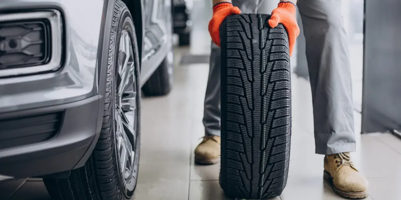 Meccanico con guanti arancioni tiene un nuovo pneumatico, pronto per essere montato su un'auto grigia. La durata gomme auto è fondamentale per la sicurezza.