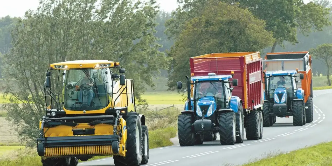 Tre trattori, uno giallo e due blu con rimorchi, percorrono una strada. Si rispettano i limiti di velocità macchine agricole.