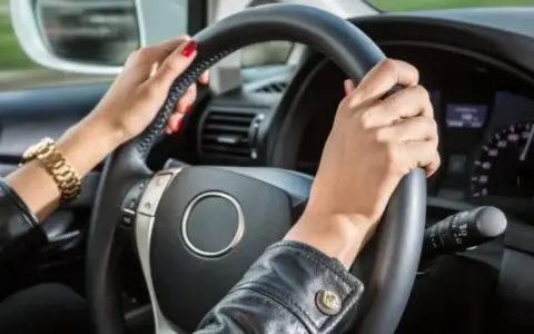 Mani con smalto rosso e bracciale d'oro tengono il volante. Il cruscotto mostra la velocità, definendo cosa vuol dire anteriore in un'auto.