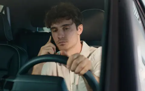 Giovane uomo al volante con il cellulare all'orecchio, distratto dalla conversazione.