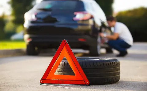 Triangolo di emergenza e ruota sgonfia accanto a un'auto nera, con un uomo che lavora alla ruota posteriore.