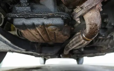 Sotto un'auto, si nota una **perdita olio motore dalla testata**, con accumuli scuri e gocciolamenti sul carter e sul tubo di scarico.