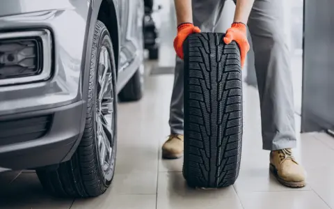 Meccanico con guanti arancioni tiene un nuovo pneumatico, pronto per essere montato su un'auto grigia. La durata gomme auto è fondamentale per la sicurezza.