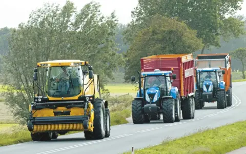 Tre trattori, uno giallo e due blu con rimorchi, percorrono una strada. Si rispettano i limiti di velocità macchine agricole.