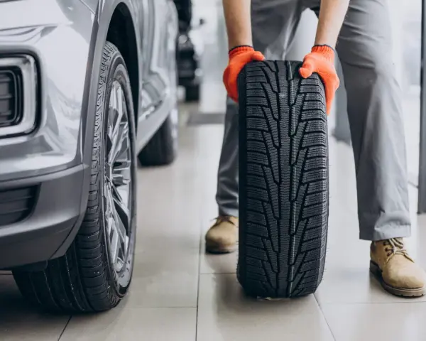 Meccanico con guanti arancioni tiene un nuovo pneumatico, pronto per essere montato su un'auto grigia. La durata gomme auto è fondamentale per la sicurezza.