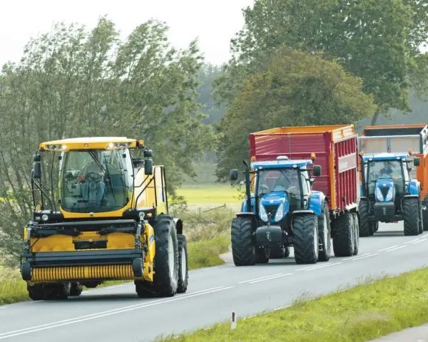 Tre trattori, uno giallo e due blu con rimorchi, percorrono una strada. Si rispettano i limiti di velocità macchine agricole.