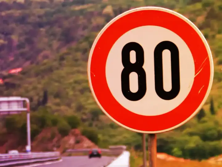 Segnale di limite massimo nei centri abitati: 80 km/h. Strada panoramica con vegetazione sullo sfondo.
