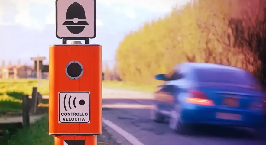 Autovelox arancione sul ciglio della strada, pronto a multare chi sfora la velocità. Un'auto blu sfreccia via, ignara.