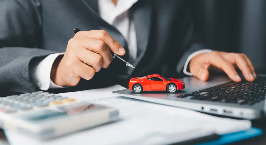 Persona in giacca grigia analizza costi auto con calcolatrice e laptop, accanto a un modellino rosso. Si chiede ogni quanto si paga il bollo auto.