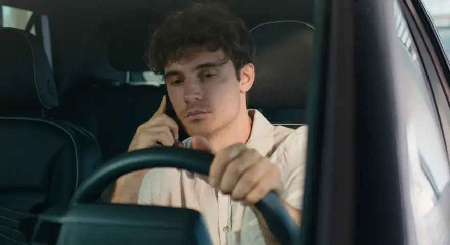Giovane uomo al volante con il cellulare all'orecchio, distratto dalla conversazione.