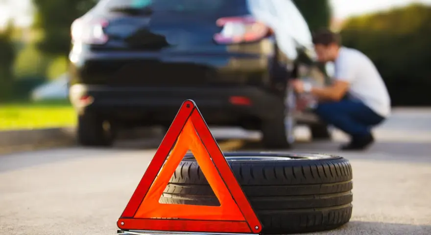 Triangolo di emergenza e ruota sgonfia accanto a un'auto nera, con un uomo che lavora alla ruota posteriore.