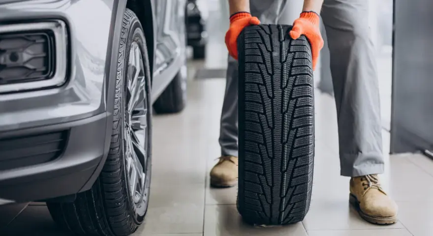 Meccanico con guanti arancioni tiene un nuovo pneumatico, pronto per essere montato su un'auto grigia. La durata gomme auto è fondamentale per la sicurezza.