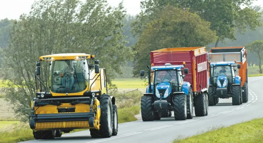 Tre trattori, uno giallo e due blu con rimorchi, percorrono una strada. Si rispettano i limiti di velocità macchine agricole.