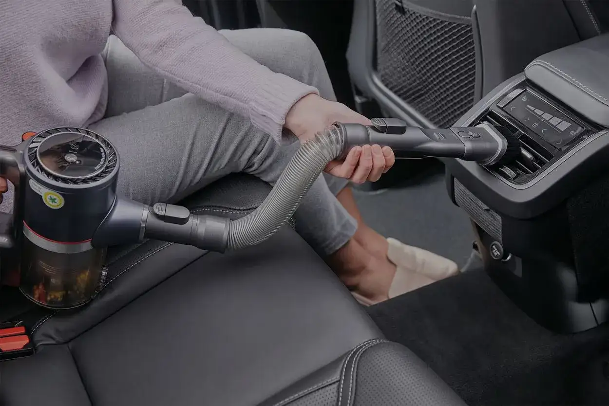 Persona pulisce l'interno dell'auto con un aspirapolvere portatile, raggiungendo anche le bocchette dell'aria per pulire vomito o sporco.