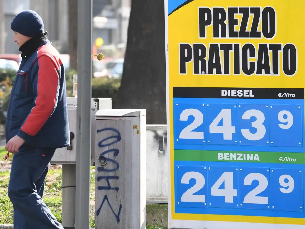 Prezzo praticato per la benzina: 2,429 &euro;/litro. Un uomo con abiti da lavoro osserva il cartellone.