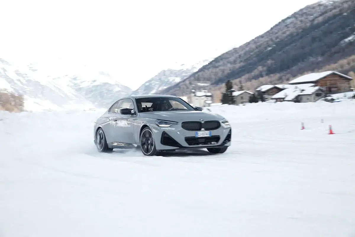 Auto grigia in derapata su pista innevata, con montagne e case sullo sfondo. Impara come guidare sulla neve con sicurezza.