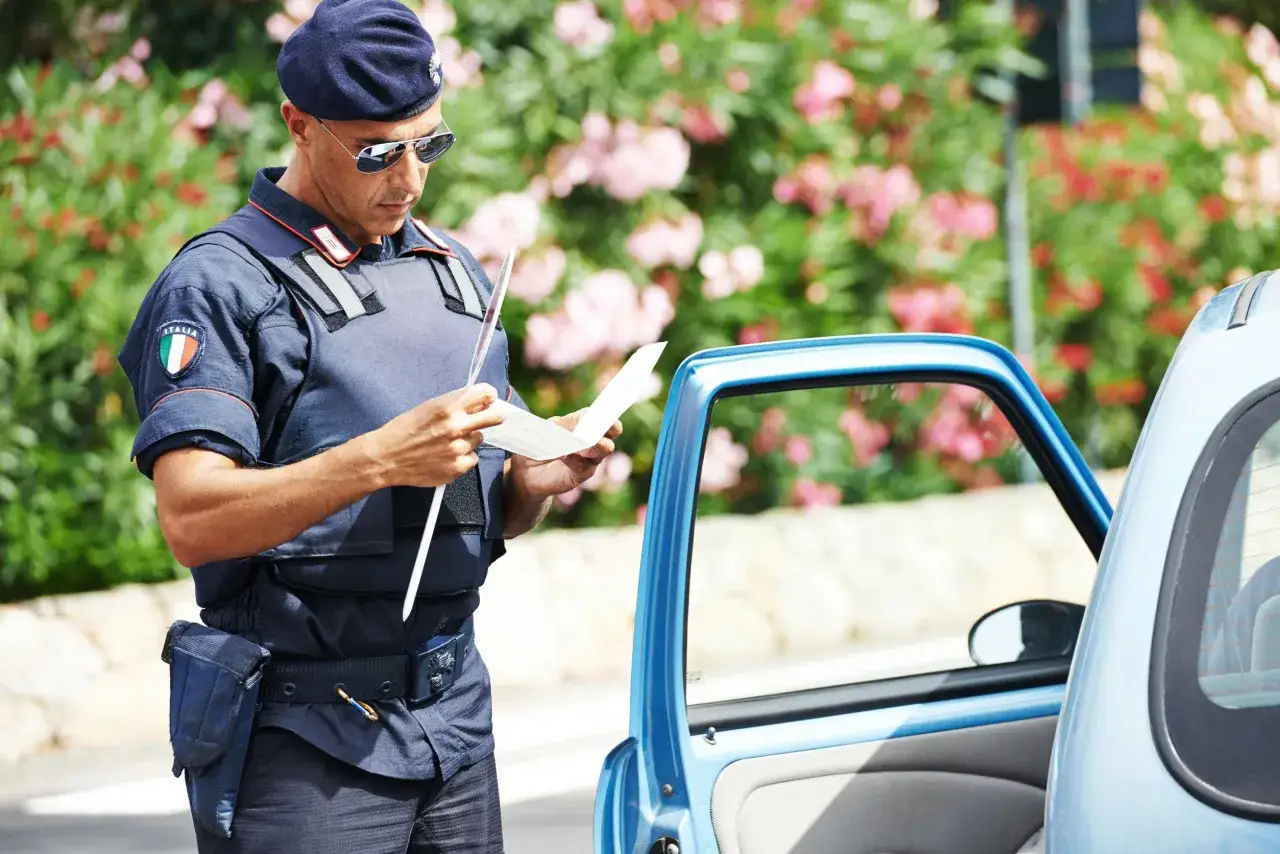 Agente di polizia italiano controlla i documenti obbligatori in auto, con un'auto blu e fiori sullo sfondo.